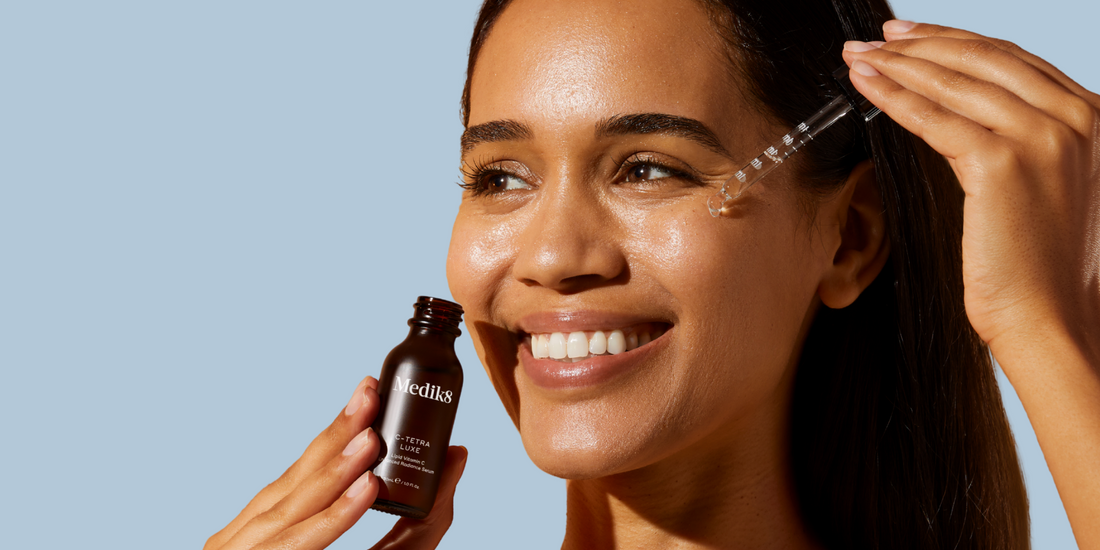 Smiling woman applying Medik8 C-Tetra Luxe Vitamin C serum to her face, promoting brightening, collagen support, and antioxidant protection.